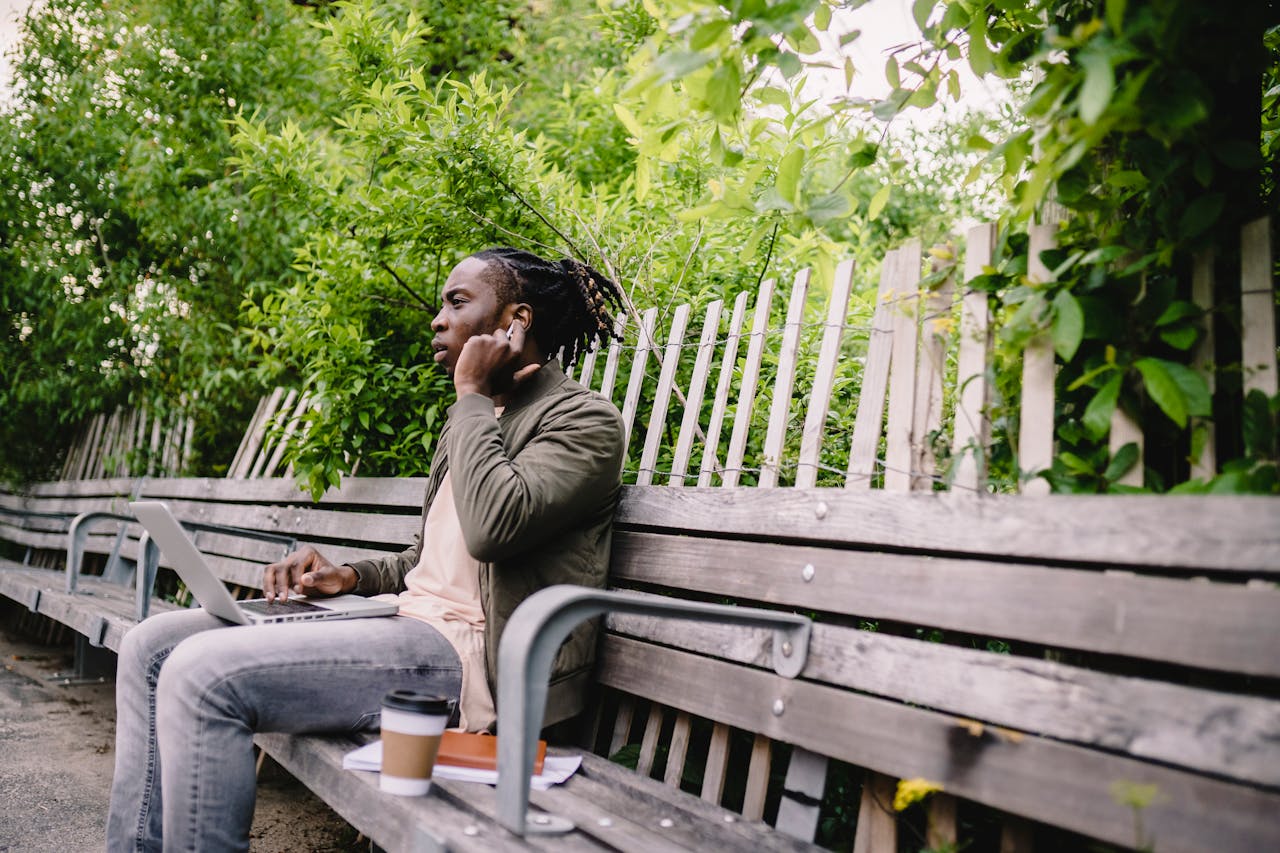 Young man working outdoors on a laptop, using earbuds, seated on a park bench.